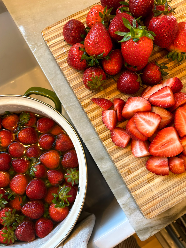 red ripe strawberries being cut on a wooden cutting board next to a colander of freshly washed berries in a sink