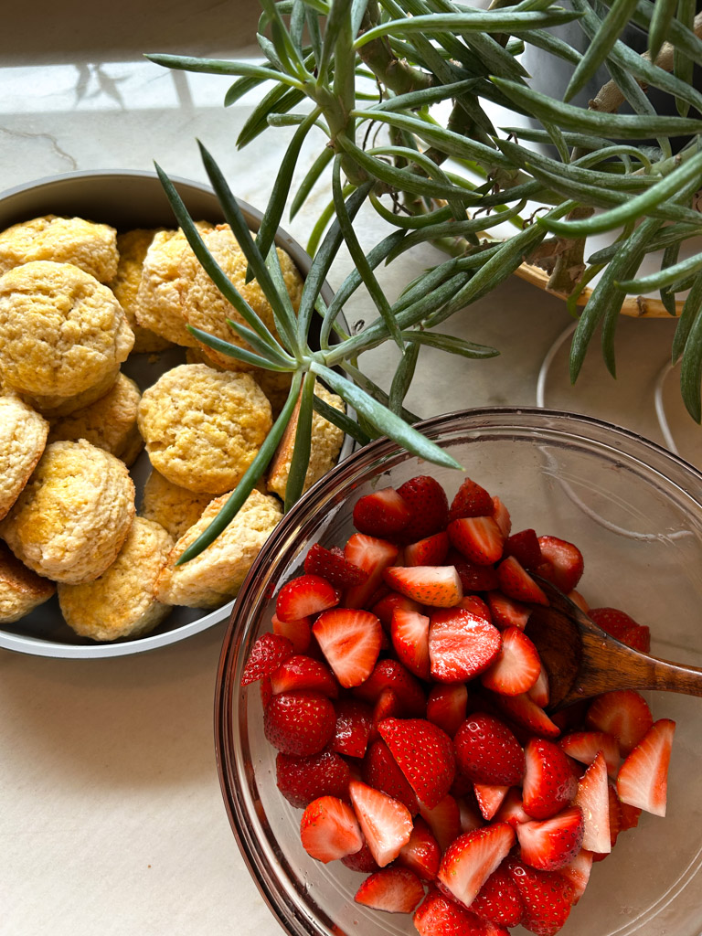 bowls of ripe red strawberries and golden brown biscuits sit on a counter next to a green plant