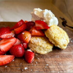Fresh strawberries and whipped cream piled on top of a shortcake biscuit sitting on a wooden cutting board