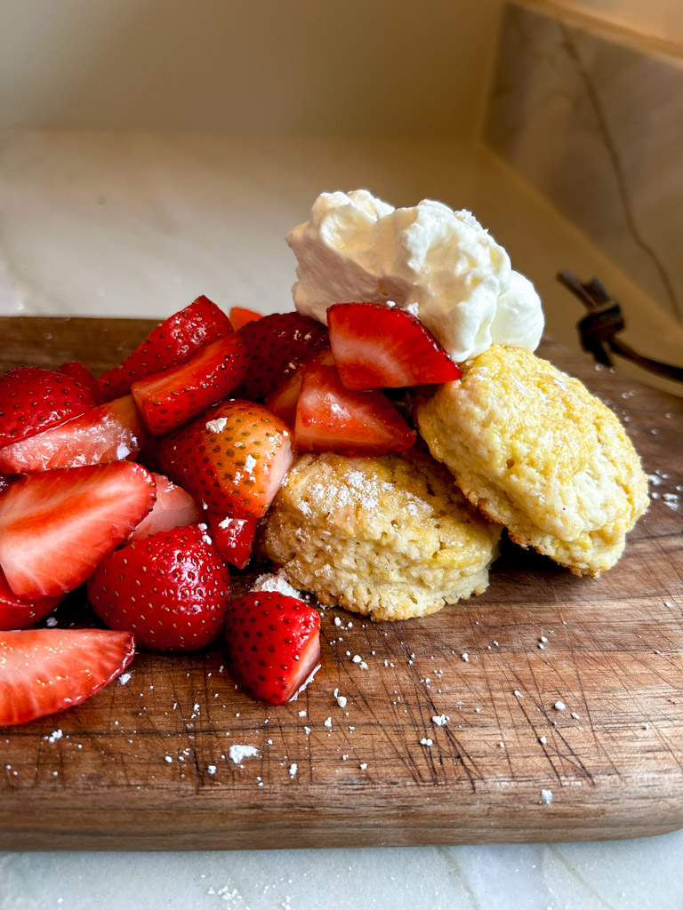 Fresh strawberries and whipped cream piled on top of a shortcake biscuit sitting on a wooden cutting board