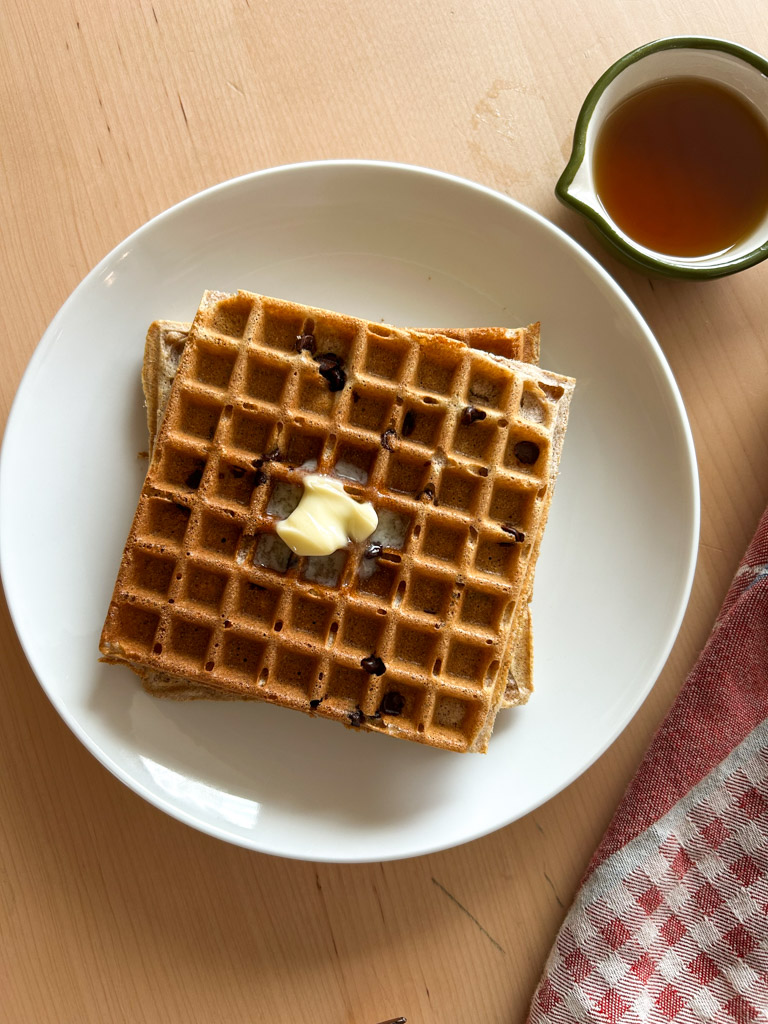 plate of buttered blegian spelt waffles sitting on the counter along with syrup cup and a tea towel
