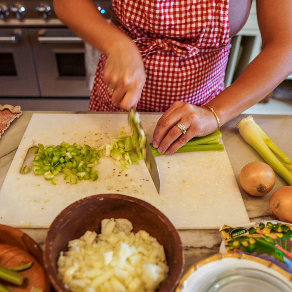 Woman wearing a red apron chops celery and onions with other fresh ingredients sitting around her on the counter