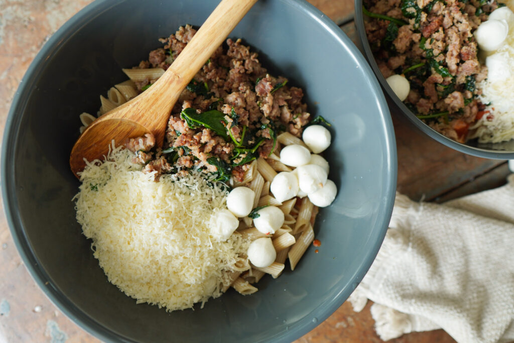 Browned sausage, spinach, pasta, and cheeses fill a blue mixing bowl and wooden spoon sitting on a wooden counter top and white tea towel