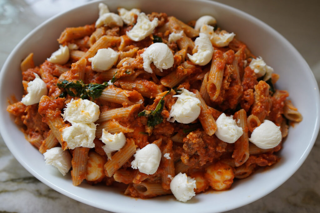 White dish filled with sausage pasta bake sits on a wooden counter top along with a tea towel
