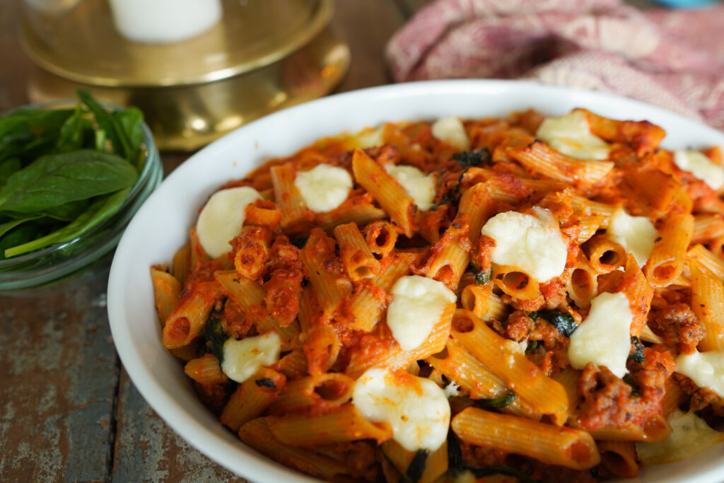 White dish filled with sausage pasta bake sits on a wooden counter top along with a tea towel