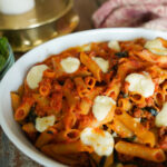 White dish filled with sausage pasta bake sits on a wooden counter top along with a tea towel