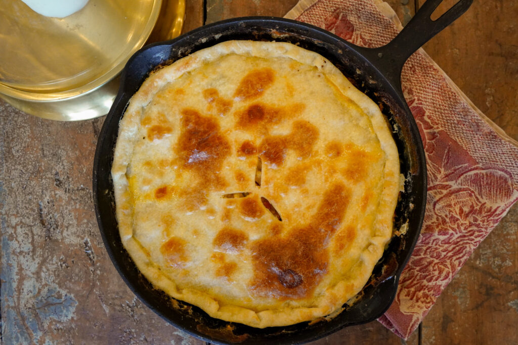 Golden crusted chicken pot pie baked to perfection in a large cast iron skillet sits on a tea towel on a wooden table top