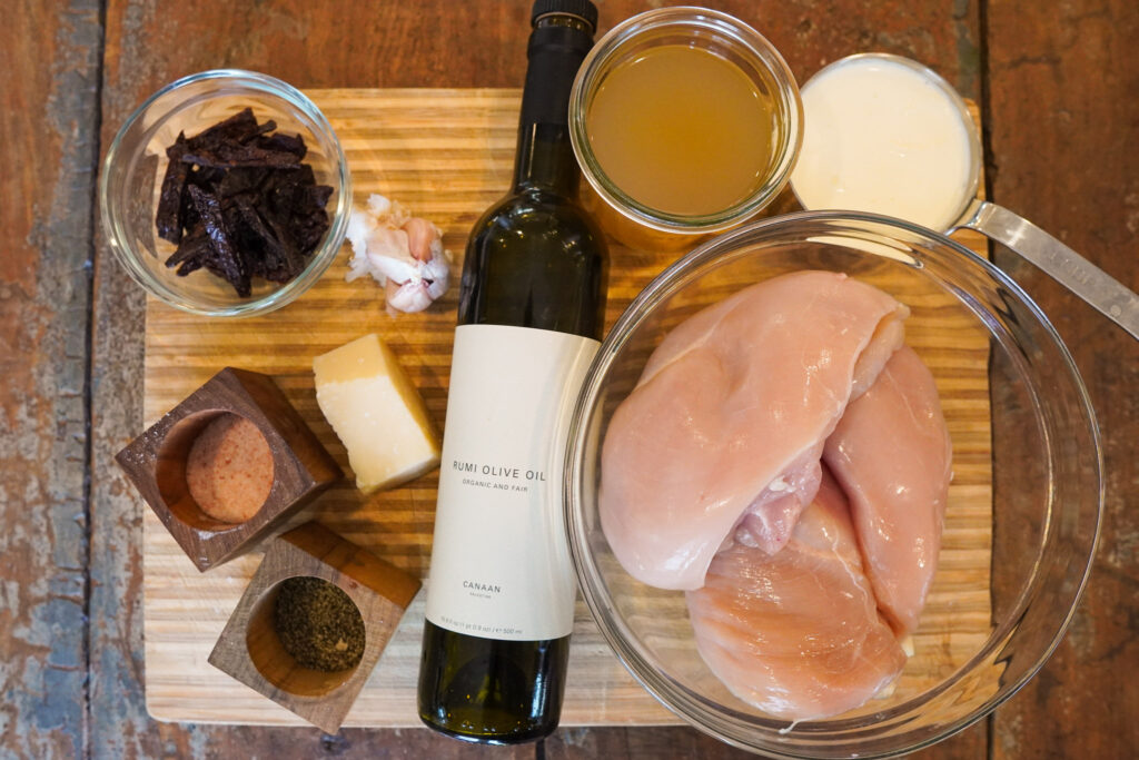 ingredients for tuscan chicken sitting on a wooden butcher block including chicken, olive oil, sun dried tomatoes, cream, and broth
