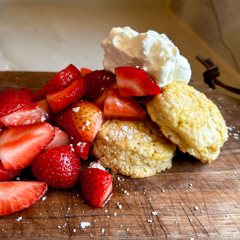 Fresh strawberries and whipped cream piled on top of a shortcake biscuit sitting on a wooden cutting board