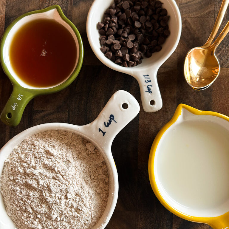 measuring cups full of flour, milk, chocolate chips, and maple syrup sitting on a wooden butcher block along with a gold mixing spoon