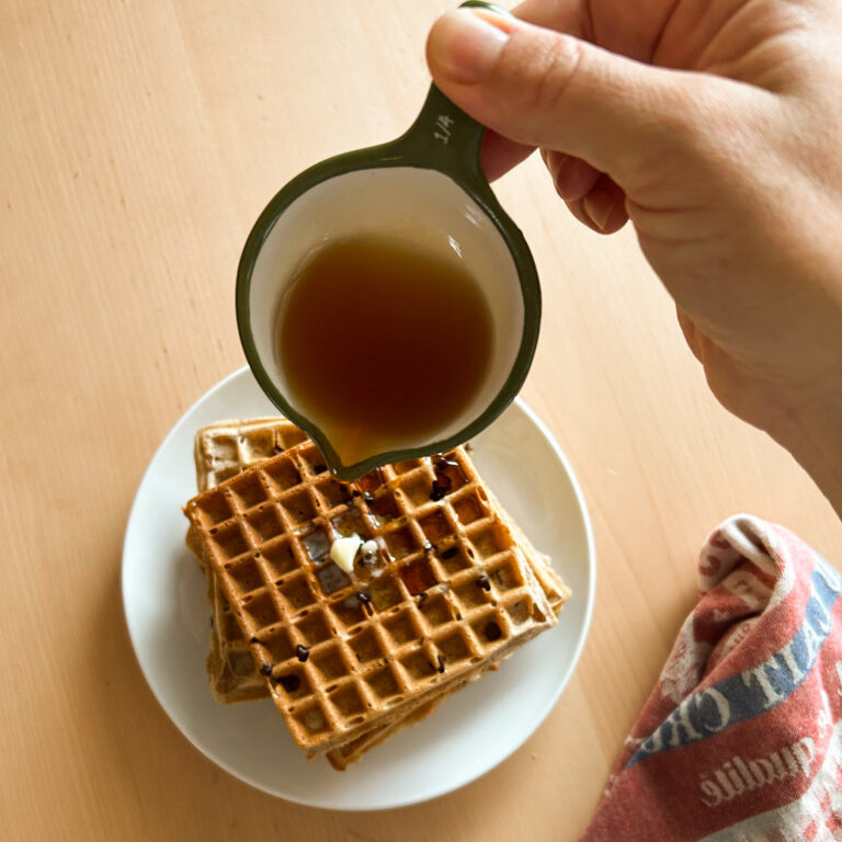 pouring cup of syrup over plate of buttered Belgian spelt waffles