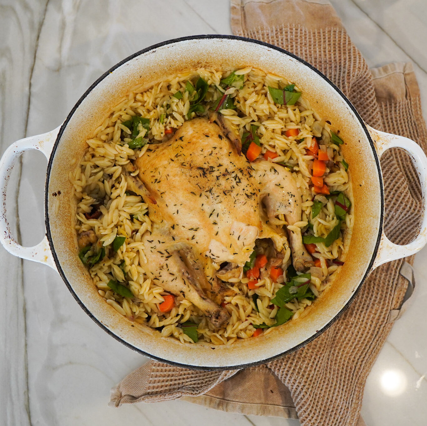 A large white dutch oven pan filled with lemon chicken and orzo sits on a white counter top along with a tea towel