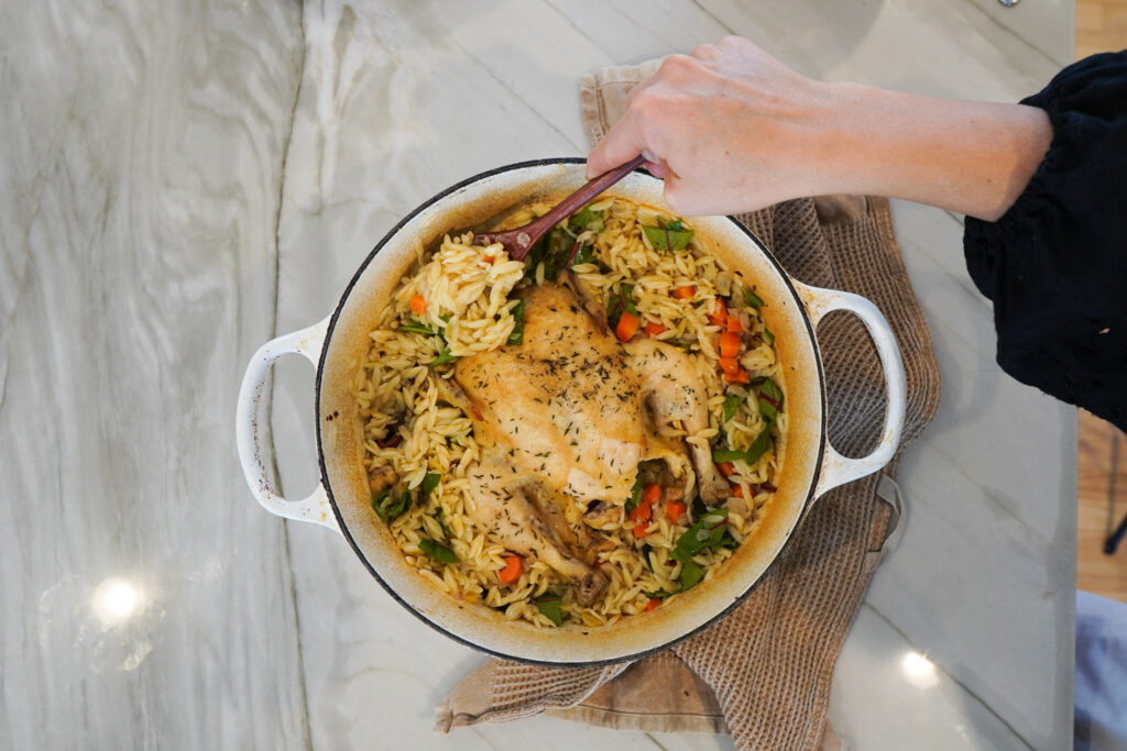 A large white dutch oven pan filled with lemon chicken and orzo sits on a white counter top along with a tea towel while someone scoops a serving of orzo from the pan