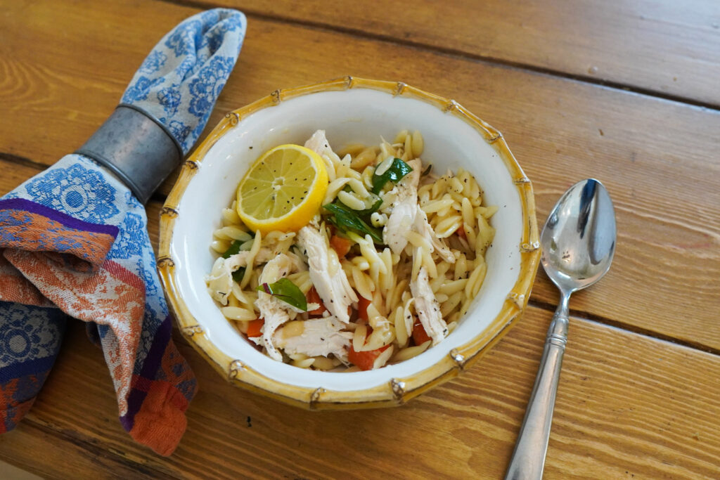 A white bowl of lemon chicken and orzo sits on a wooden table top with a cloth napkin and spoon