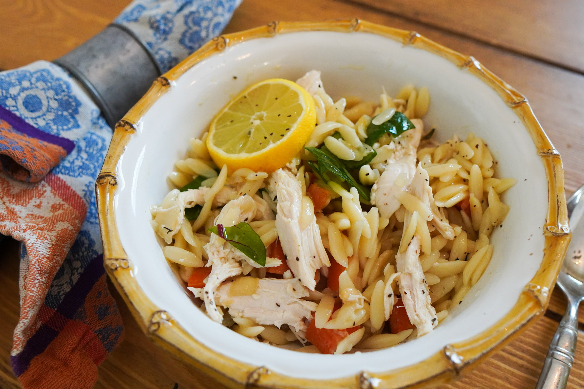 A white bowl of lemon chicken and orzo sits on a wooden table top with a cloth napkin and spoon