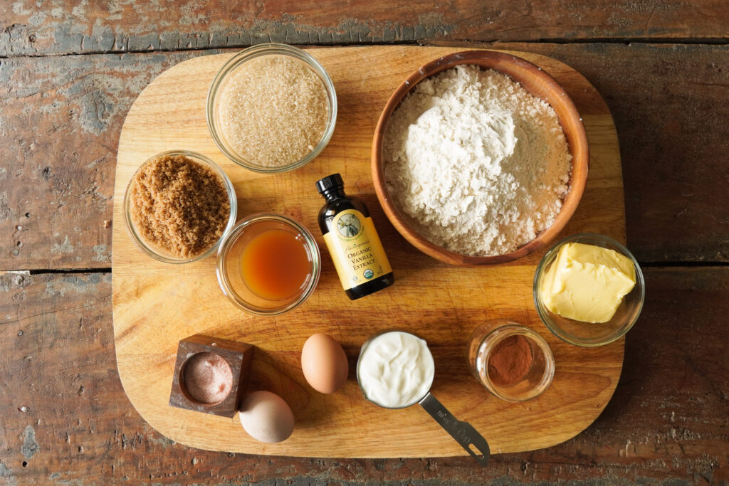 Ingredients for an apple cider spice cake sit on a wooden cutting board including flour, sugar, eggs, butter, vanilla, and apple cider