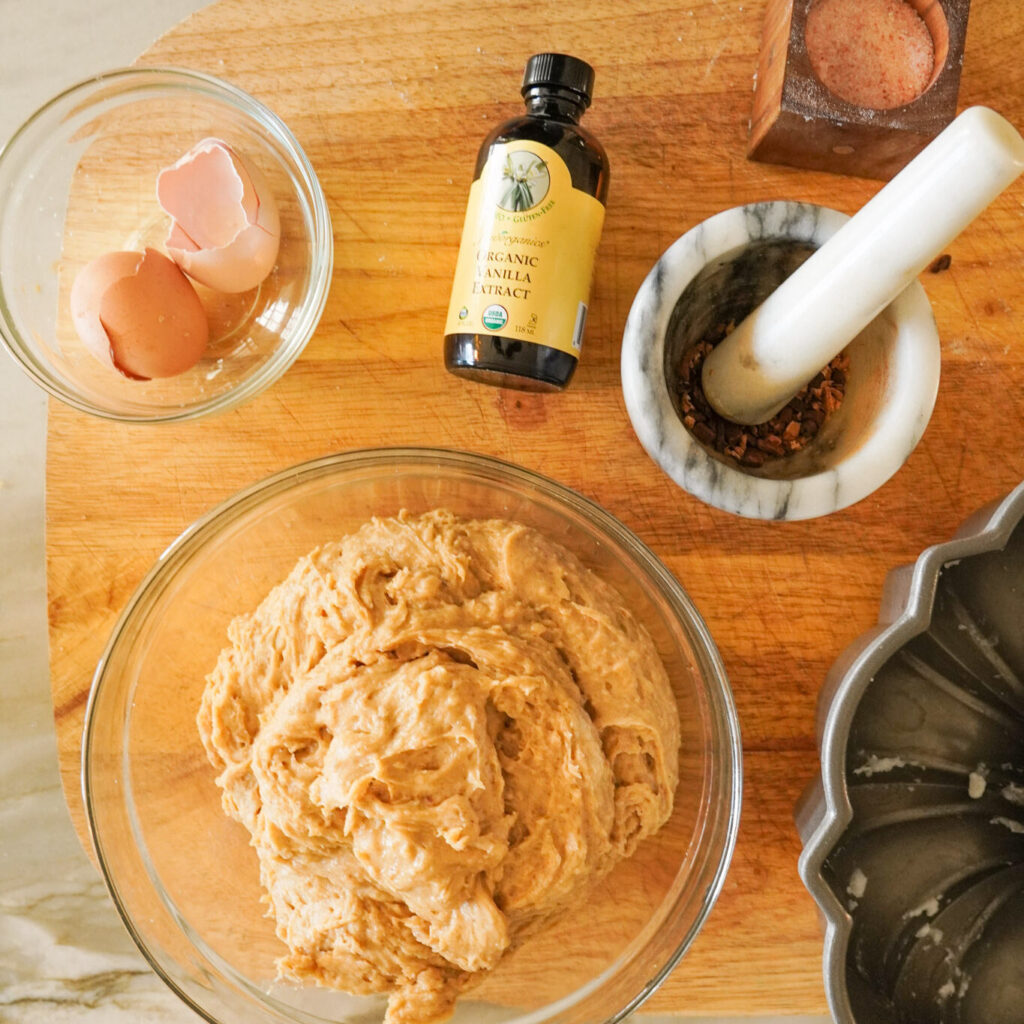 A bowl of cake batter and other various ingredients for apple cider spice cake sit on a wooden cutting board