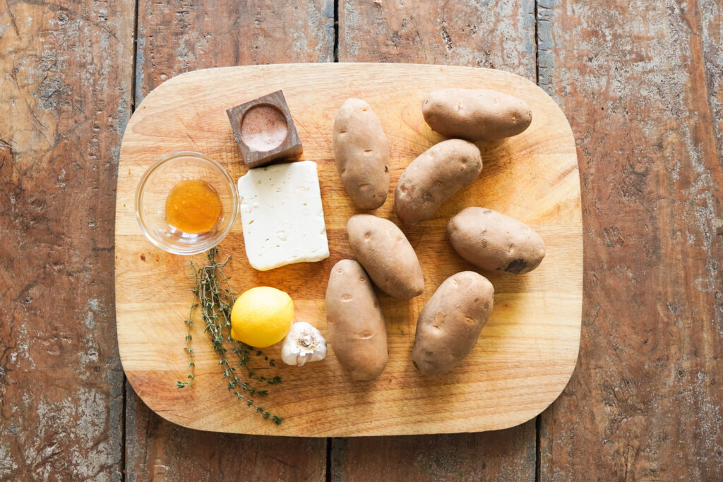 Ingredients for crispy feta potatoes sitting on a wooden cutting board including potatoes, feta cheese, lemon and garlic