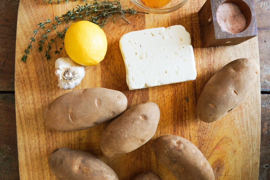 Ingredients for crispy feta potatoes sitting on a wooden cutting board including potatoes, feta cheese, lemon and garlic