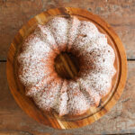 An apple cider spice cake dusted with powdered sugar sits on a wooden cake platter on a table top