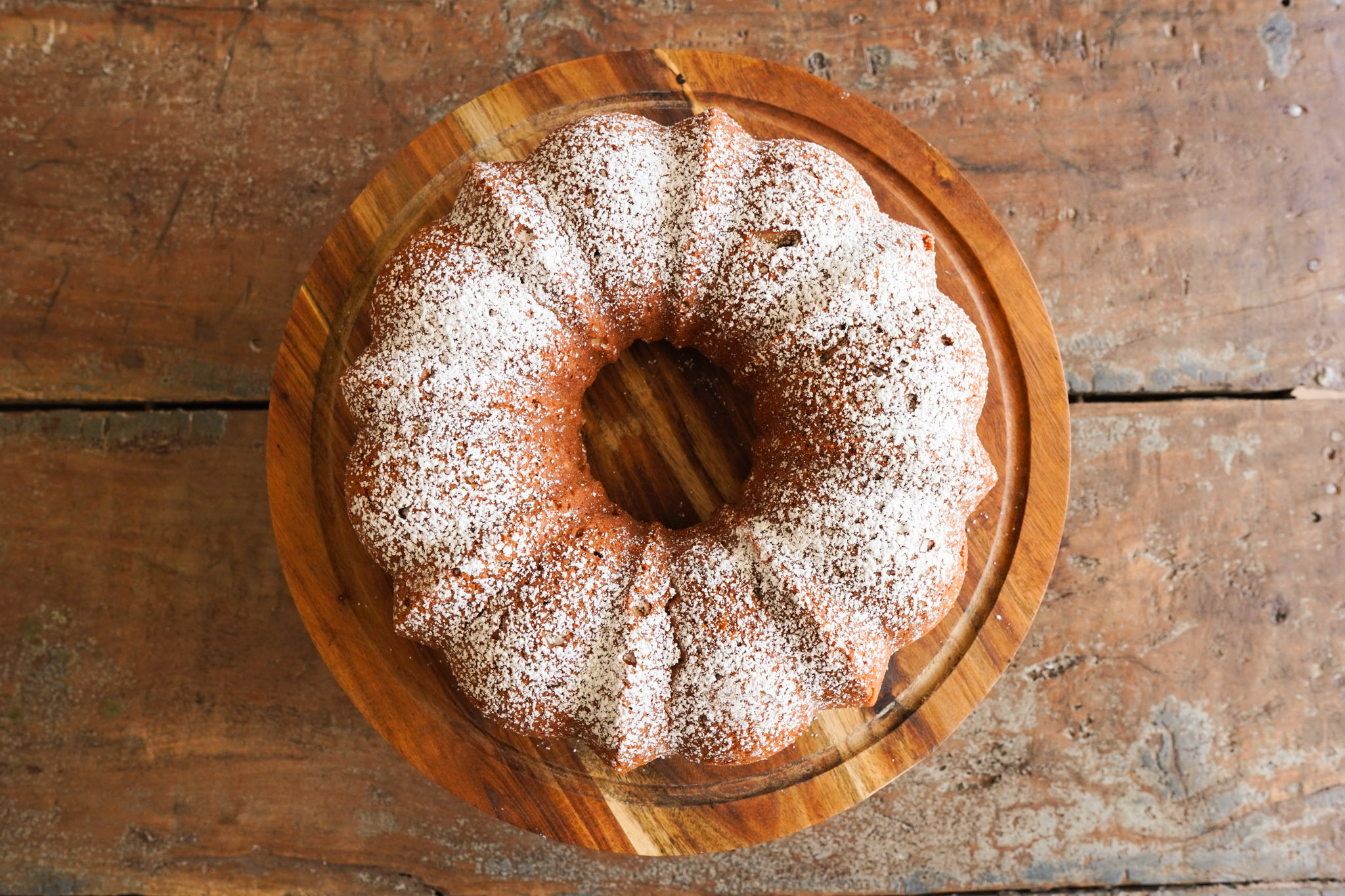 An apple cider spice cake dusted with powdered sugar sits on a wooden cake platter on a table top