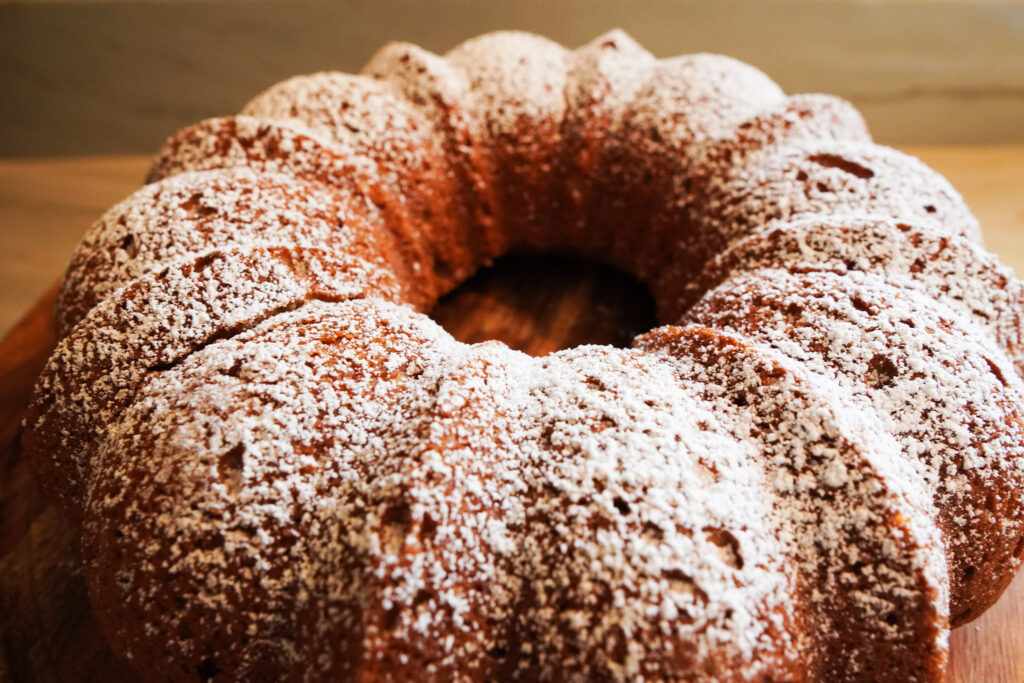 An apple cider spice cake dusted with powdered sugar sits on a wooden cake platter on a table top