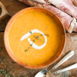 wooden bowl of cozy butternut squash soup sitting on a wooden table along with spoons and a tea towel