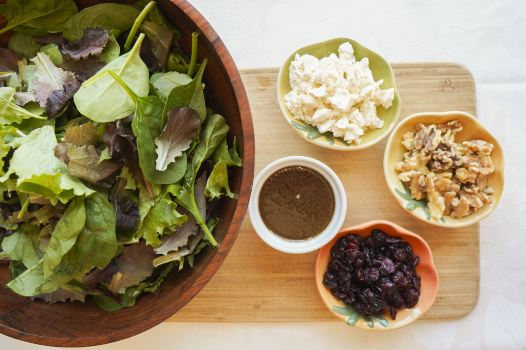Bowls of ingredients for winter salad sitting on a wooden cutting board including greens, walnuts, cranberries, and feta