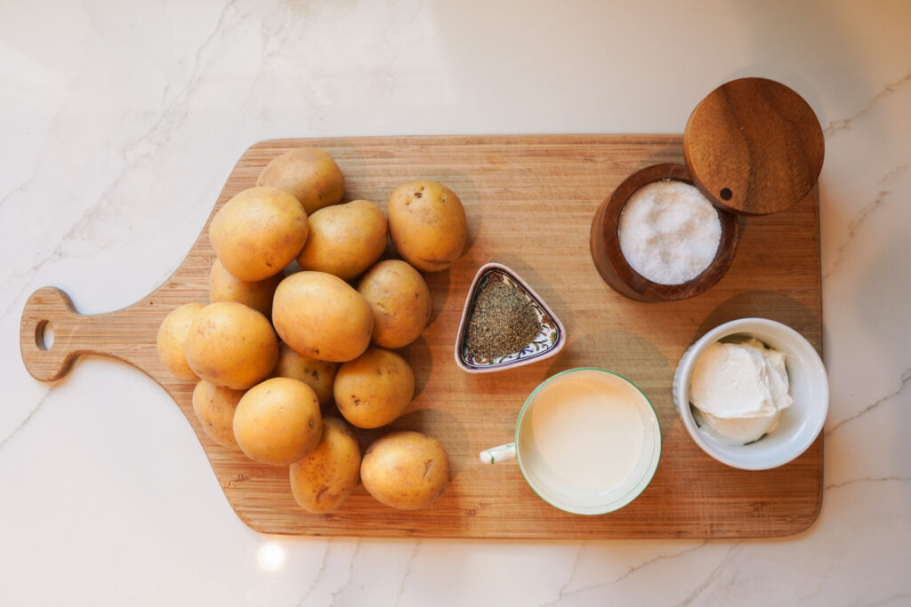 Ingredients for creamy mashed potatoes sits on a wooden cutting board including yukon gold potatoes, cream, salt, and sour cream