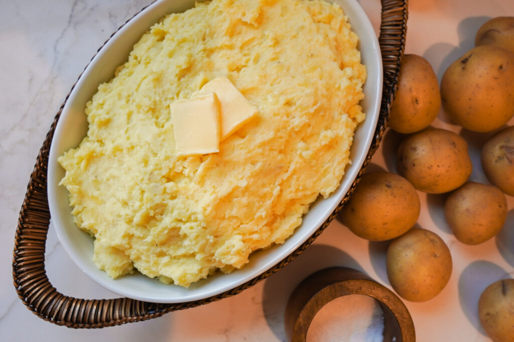 A white casserole dish filled with creamy mashed potatoes sits on the counter next to a pile of whole yukon gold potatoes and salt