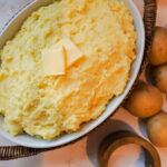 A white casserole dish filled with creamy mashed potatoes sits on the counter next to a pile of whole yukon gold potatoes and salt