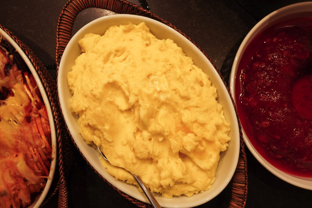 A family dinner table is filled with hearty dishes including mashed potatoes, sweet potatoes, and cranberry sauce